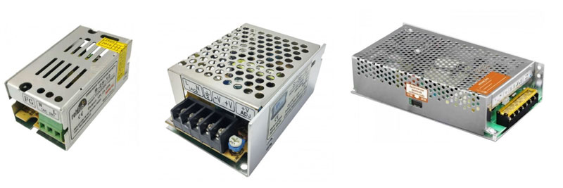 rata-se de uma imagem mostrando três fontes de alimentação chaveadas (switching power supplies) industriais, em invólucros metálicos retangulares e ventilados. Elas possuem terminais de parafuso para conexão de entrada AC e saída DC (tensão contínua), usadas para alimentar eletrônicos