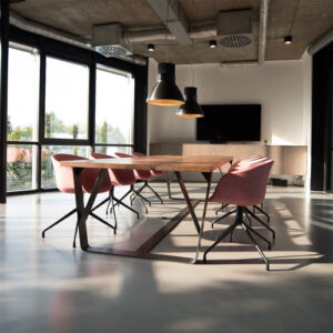 Um moderno escritório com teto e tubulações aparentes. Uma longa mesa de reunião de madeira clara, com pés metálicos escuros em "X", está rodeada por cadeiras giratórias rosa salmão. Grandes janelas trazem muita luz natural. Duas luminárias pendentes pretas e largas pairam sobre a mesa.