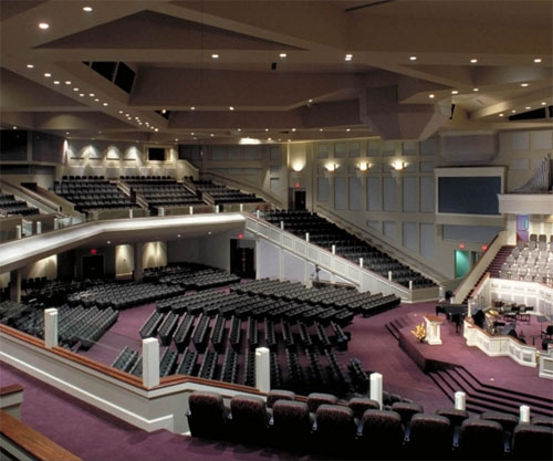  Uma vista interna ampla de um grande auditório de igreja, moderno e com arquitetura angular. Possui muitos assentos escuros em degraus e carpete roxo. O palco e o púlpito estão à direita. O ambiente é iluminado, com luzes embutidas no teto e paredes.