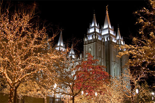 Essa é uma foto noturna de um grande templo, provavelmente o Templo de Salt Lake, com pináculos brancos iluminados. Em primeiro plano, árvores estão cobertas por milhares de pequenas luzes brancas e douradas, criando um brilho festivo e quente, contrastando com o céu escuro da noite. Parece ser inverno, com um pouco de neve nas árvores e no topo do templo