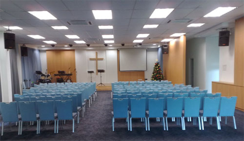 A imagem mostra o interior de um salão de reuniões, possivelmente uma igreja moderna. Há fileiras de cadeiras azuis voltadas para um palco simples. No palco, há um púlpito, um teclado e, ao fundo, uma cruz de madeira e uma tela de projeção. O teto tem iluminação embutida.