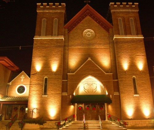 A imagem noturna mostra a fachada de uma igreja ou templo de tijolos. O edifício tem duas torres laterais e uma entrada central em arco. A iluminação de baixo para cima destaca a textura dos tijolos e os elementos arquitetônicos, criando um efeito dramático e acolhedor. Há enfeites de Natal na entrada, com guirlandas e laços vermelhos.