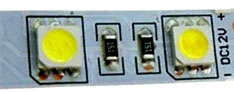 Uma tira de LED flexível branca. Há dois LEDs quadrados amarelos nas pontas e dois pequenos resistores pretos no centro. No lado direito, vê-se o texto "DC12V +", indicando a voltagem. O fundo é de um circuito impresso claro com trilhas metálicas discretas.