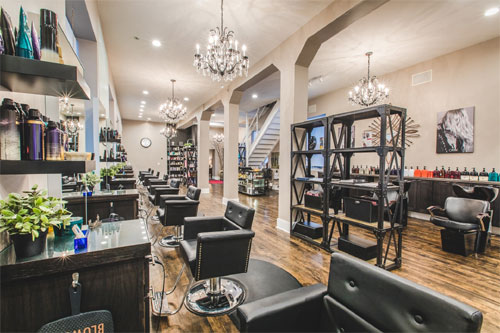 O piso é de madeira escura e polida. Há várias estações de cabeleireiro alinhadas, cada uma com uma cadeira de couro preta e espelho. No centro, uma prateleira metálica escura exibe produtos. O teto alto possui lustres de cristal. Uma escada no fundo leva a um segundo andar. O ambiente é sofisticado, com paredes em tons neutros e iluminação suave.