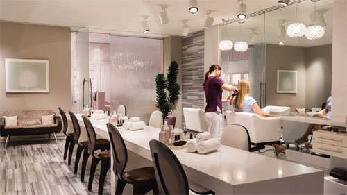 O piso é de madeira escura e polida. Há várias estações de cabeleireiro alinhadas, cada uma com uma cadeira de couro preta e espelho. No centro, uma prateleira metálica escura exibe produtos. O teto alto possui lustres de cristal. Uma escada no fundo leva a um segundo andar. O ambiente é sofisticado, com paredes em tons neutros e iluminação suave