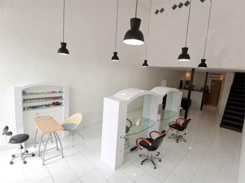  A imagem mostra o interior moderno e bem iluminado de um salão de beleza. O piso e as paredes são brancos. No centro, há duas estações de cabeleireiro com espelhos curvos e cadeiras pretas e laranja. À esquerda, uma área de manicure com prateleiras cheias de esmaltes. O teto alto tem luminárias pendentes pretas. No canto direito superior, há uma escada escura que leva a outro andar.