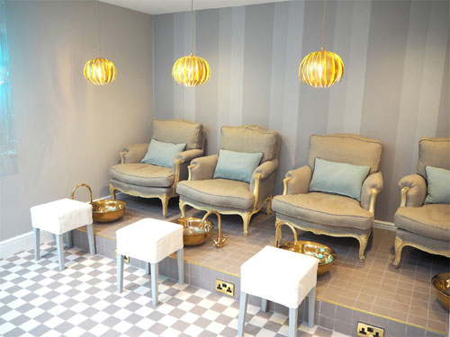  Esta é uma sala de pedicure luxuosa e elegante. Há quatro poltronas individuais estofadas em tom bege, com detalhes dourados na madeira, dispostas em uma plataforma elevada. Em frente a cada uma, no chão, há um banquinho branco para o cliente e uma bacia dourada para os pés. O chão é xadrez cinza e branco. As paredes são cinza claro com listras verticais mais escuras, e três luminárias pendentes douradas iluminam o ambiente.