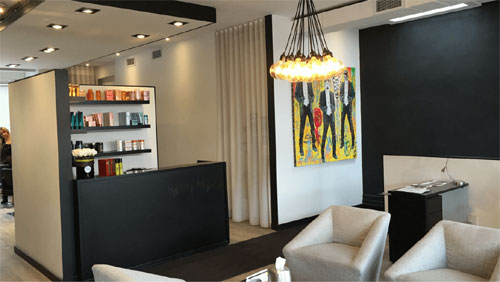  Este é o interior moderno de um salão de beleza. A recepção é um balcão preto com prateleiras cheias de produtos ao lado. Há duas poltronas estofadas claras para espera e uma mesinha lateral. Uma pintura colorida e abstrata com figuras humanas se destaca na parede escura. A iluminação é feita por um lustre pendente com várias lâmpadas. O ambiente tem paredes em branco e preto, com piso de madeira clara.