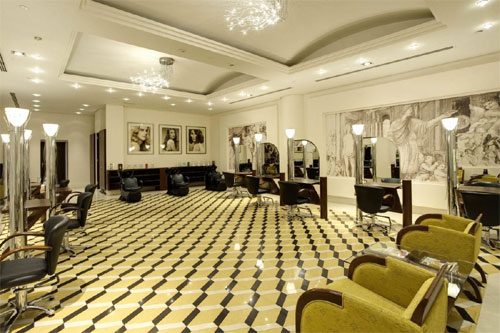  Este é o interior luxuoso de um salão de beleza. O piso tem um padrão geométrico chamativo em amarelo e preto. Há várias estações de cabeleireiro com cadeiras de couro pretas e espelhos grandes. A iluminação é sofisticada, com lustres no teto e luminárias de coluna. As paredes são claras, com obras de arte e fotos de modelos. O ambiente é espaçoso e elegante.