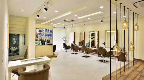  Esta é a imagem de um salão de beleza moderno e sofisticado. O ambiente é claro, com paredes em tons neutros. No centro, há várias estações de trabalho, cada uma com uma poltrona de couro marrom e um espelho grande e iluminado. No canto direito, uma divisória decorativa é feita de hastes pretas verticais com pendentes de vidro em formato de gota. Há também uma área de recepção ou manicure em primeiro plano. A iluminação é composta por spots e luzes embutidas.