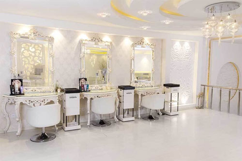 Uma sala de salão de beleza elegante, predominantemente branca e dourada, com estilo clássico/barroco. Possui três estações de trabalho simétricas, cada uma com uma cadeira estofada branca, uma penteadeira ornamentada de pernas curvas e um espelho grande com moldura elaborada. O piso é claro e liso. A iluminação é suave e o teto tem detalhes em gesso e luminárias pendentes sofisticadas. O ambiente é luxuoso e organizado.