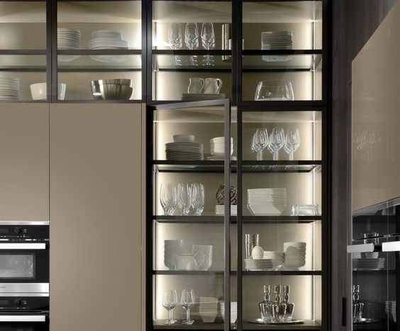 A foto mostra um armário de cozinha ou cristaleira com portas de vidro e estrutura escura. No interior, há prateleiras de vidro repletas de louças brancas (pratos, xícaras) e taças/copos de vidro transparente. O grande destaque é a iluminação por fita de LED instalada nas laterais e sob cada prateleira 