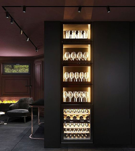  É uma foto de um móvel preto em uma sala escura. O móvel tem prateleiras de vidro iluminadas por dentro com luz amarela, exibindo taças e garrafas de vinho. O ambiente ao redor parece ser um espaço de estar aconchegante, com uma poltrona escura e uma lareira acesa visível à esquerda.