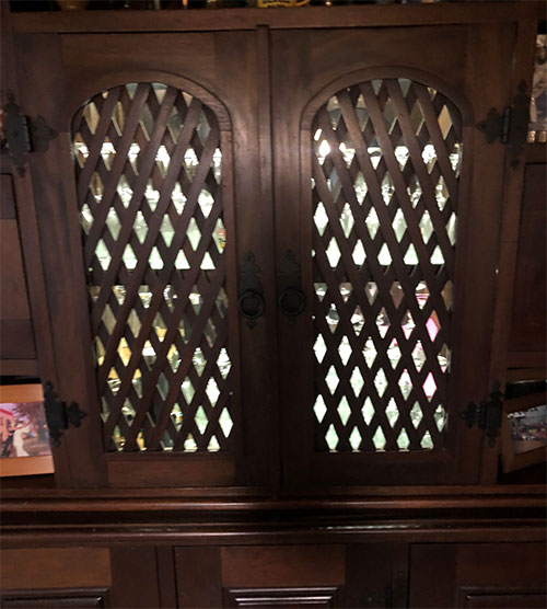 A imagem mostra a parte superior de um móvel de madeira escura, que parece ser uma cristaleira ou adega. Possui duas portas arqueadas cada uma com detalhe de treliça diagonal (losangos) vazada. Há uma luz interna brilhante de LED) que ilumina o interior e se projeta através dos vãos da treliça