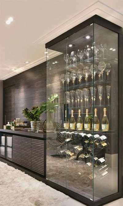 Uma cristaleira/adega moderna de vidro, iluminada por fita de LED no topo, destaca taças e garrafas de espumante. Ao lado, um longo balcão escuro com acabamento ripado e área para bebidas, criando um ambiente sofisticado.