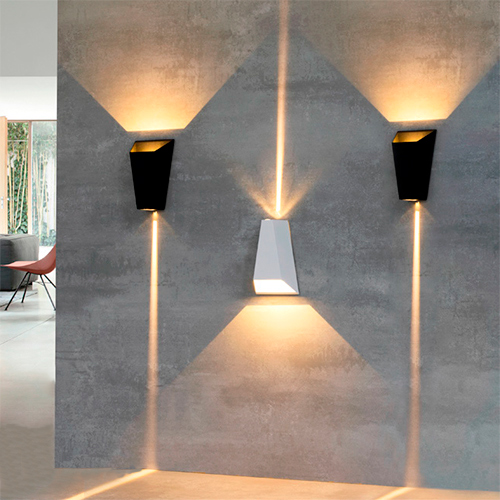 
Esta imagem mostra três arandelas modernas com design geométrico, instaladas em uma parede de cimento queimado. Elas possuem formato de trapézio e emitem luz para cima e para baixo. Os fachos criam desenhos de triângulos largos e linhas finas e intensas na parede, gerando um efeito decorativo sofisticado.