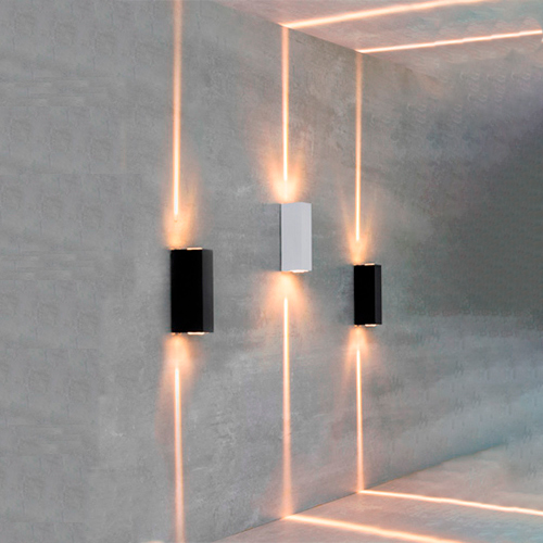 
Três luminárias retangulares fixas em uma parede cinza projetam feixes de luz verticais e finos para cima e para baixo. A luz é quente e alaranjada, criando linhas retas que se estendem até o teto e o chão, formando um desenho geométrico moderno e minimalista no ambiente.