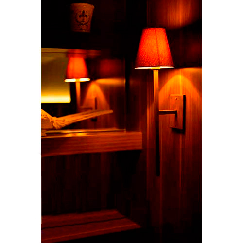 Imagem de um ambiente interno aconchegante, com paredes de madeira. Uma arandela de parede com cúpula vermelha emite luz quente e suave, criando sombras e reflexos no espelho ao fundo, transmitindo sensação de conforto e intimidade.