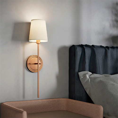 Imagem de um quarto moderno. Na parede há uma arandela com base redonda e haste vertical dourada, com cúpula clara acesa. Ao lado, vê-se a cabeceira estofada azul da cama e parte de uma poltrona clara, criando um ambiente aconchegante.