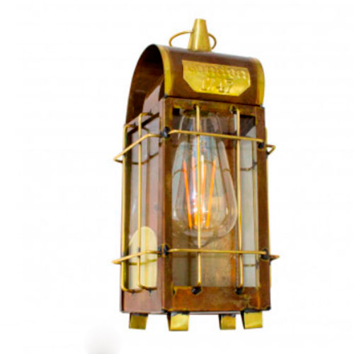 Lanterna rústica de parede em metal cobre e detalhes dourados. Possui formato retangular com teto curvado, protegida por uma grade de metal. No centro, brilha uma lâmpada de filamento aparente com luz alaranjada. No topo, há uma placa gravada "London 1747". Estilo vintage e náutico.