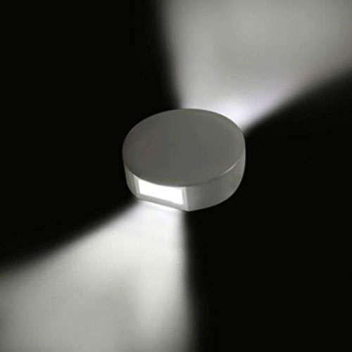 
Esta arandela branca tem formato circular e achatado, como um disco. Ela é feita de policarbonato e possui facho duplo, projetando dois triângulos de luz branca intensa em direções opostas contra a parede, criando um efeito decorativo moderno e simétrico.