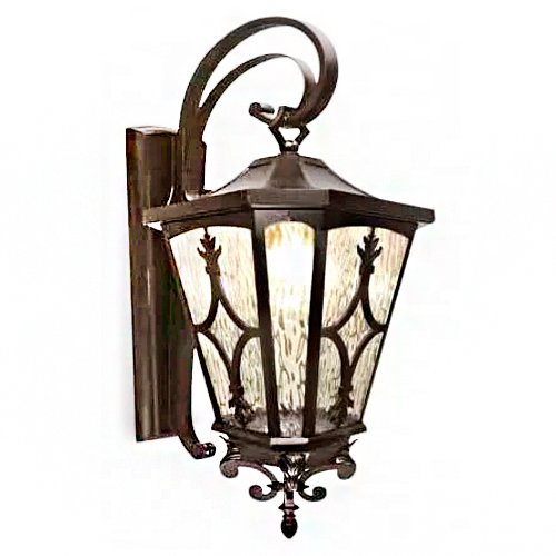 Arandela externa clássica em metal escuro. Possui um suporte de parede com um braço curvado em arco que segura uma luminária em formato de lanterna sextavada. O vidro é fosco/texturizado, decorado com arabescos e detalhes florais. Na base, há um pendente ornamental trabalhado.