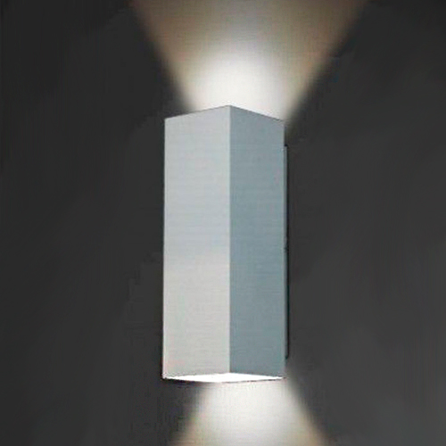 Esta arandela branca (modelo Rocca) tem formato de um retângulo vertical minimalista fixado à parede. Ela possui aberturas no topo e na base, criando dois fachos de luz em formato de triângulo que iluminam simultaneamente para cima e para baixo, destacando a textura da parede.