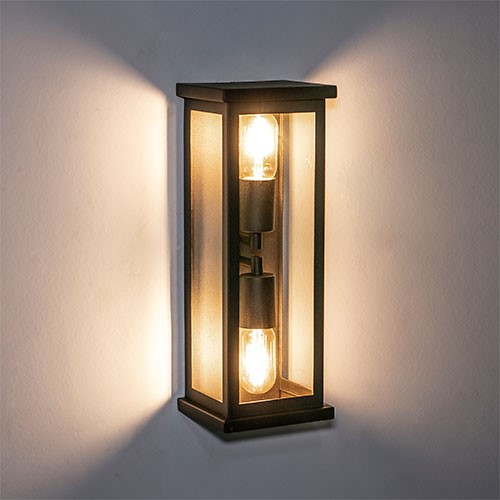 Esta arandela clássica tem formato de uma lanterna retangular vertical com estrutura metálica preta e vidros transparentes. Em seu interior, duas lâmpadas de filamento acesas criam uma iluminação acolhedora, projetando feixes de luz amarelos nas laterais da parede