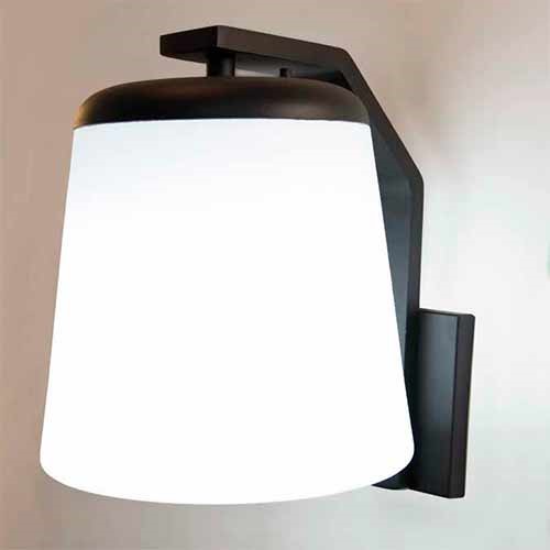 
A imagem mostra uma arandela de parede moderna. Ela possui um suporte metálico preto em formato de "L" invertido que segura uma cúpula cônica de vidro branco fosco. A luminária está acesa, emitindo um brilho suave e uniforme por toda a cúpula, contrastando com a estrutura escura e a parede clara.