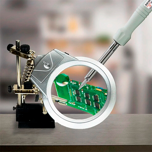 Lupa circular em um corpo prateado, que está presa a um braço ajustável de metal. Este braço se conecta a uma base preta e pesada para estabilidade. O conjunto inclui também uma ou mais garras tipo "jacaré" , que servem para segurar objetos pequenos, como componentes eletrônicos.