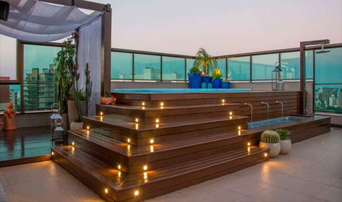 Essa imagem mostra uma área de lazer externa luxuosa, provavelmente em uma cobertura ou varanda. O destaque é uma estrutura de madeira em degraus que leva a uma piscina ou jacuzzi elevada. Os degraus têm pequenas luzes embutidas que criam um efeito aconchegante. A área tem plantas em vasos, uma cerca de vidro e, ao fundo, a vista da cidade.