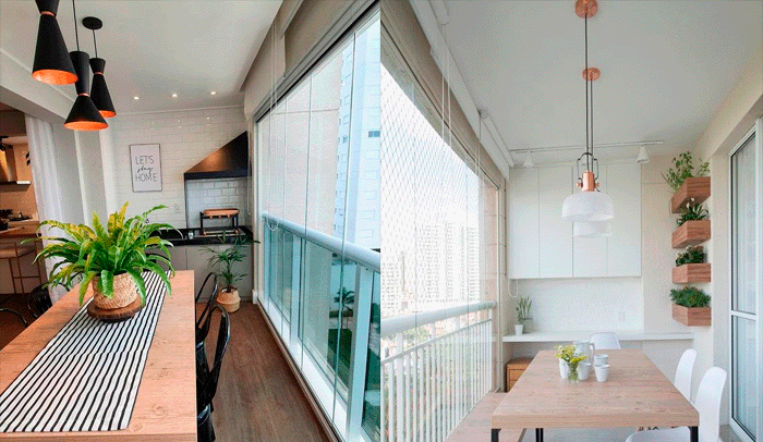 Esquerda: Varanda estreita com uma mesa de madeira clara e um caminho de mesa listrado. Há três pendentes pretos e dourados sobre a mesa. A parede tem azulejos brancos tipo 'subway' e uma churrasqueira escura embutida. O piso é de madeira escura. Há plantas, incluindo uma grande samambaia. O fechamento é de vidro, com uma tela de proteção. Direita: Varanda clara, também com mesa de madeira clara e cadeiras brancas. O teto branco tem trilhos de luz e um pendente moderno com detalhe em cobre. Na parede branca, há nichos de madeira com plantas. O ambiente é minimalista.