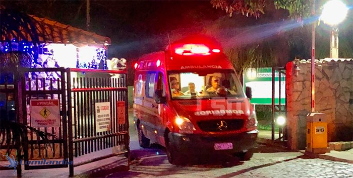 A foto noturna mostra uma ambulância vermelha com as luzes de emergência ligadas entrando em um portão, que parece ser a entrada de um condomínio. No para-brisa da van, há motorista e, pelo menos, outros dois socorristas. O portão tem luzes decorativas e placas de aviso. A cena indica uma emergência médica.