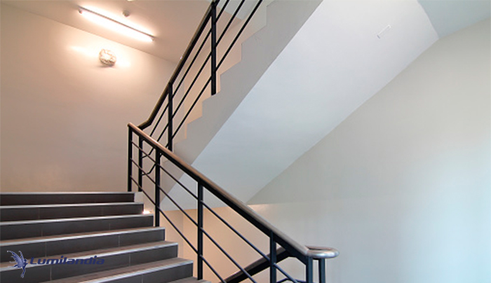 Uma escada interna com degraus de cor escura (provavelmente cinza ou marrom escuro) e um corrimão de metal preto. A escada sobe em curva, à esquerda, levando a um andar superior. As paredes são claras, em tom bege suave, com iluminação de luz fluorescente vinda do teto.