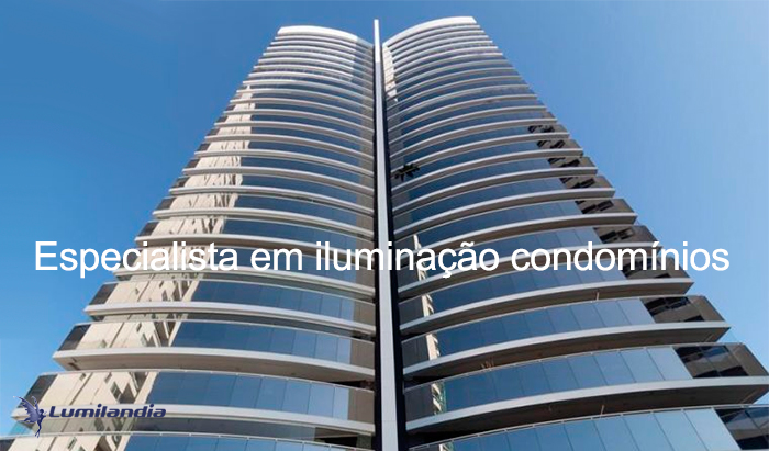 A imagem mostra a entrada de um prédio residencial alto. Há um muro baixo com portão de madeira vertical e uma portaria com grandes janelas de vidro. O paisagismo é tropical, com palmeiras e canteiros de flores coloridas ao redor de um caminho pavimentado. O dia é ensolarado, com céu azul claros 