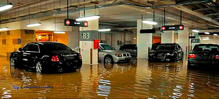 A imagem mostra uma garagem de estacionamento subterrânea completamente alagada. A água suja, em tom marrom, cobre o chão e reflete as luzes do teto. Há vários carros de luxo estacionados, com a água atingindo a altura das rodas ou um pouco acima. Sinais de "NO ENTRY" (Não entre) estão visíveis