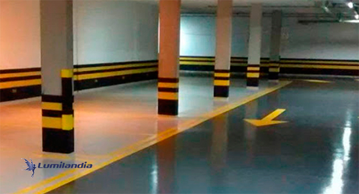 O piso é brilhante, predominantemente cinza claro, com marcações de segurança em amarelo forte. Uma seta direcional amarela é visível no centro-direita. As colunas de sustentação e a parte inferior das paredes são sinalizadas com faixas horizontais amarelas e pretas alternadas, indicando uma área de perigo 