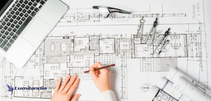 A imagem mostra a visão superior de uma planta arquitetônica ou projeto de engenharia em papel grande, onde uma pessoa está desenhando ou revisando com um lápis. No canto superior esquerdo, há um laptop aberto. Óculos e um compasso de desenho estão perto do centro superior. À direita, rolos de papel de projeto.