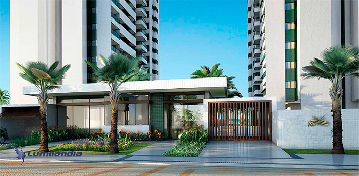 entrada de um prédio residencial moderno, com dois prédios brancos visíveis. Há um portão de acesso de madeira escura e uma portaria com teto branco horizontal e grandes janelas de vidro. A área externa é ajardinada, com grama verde, palmeiras altas nas laterais e caminhos de pedra ou azulejo.