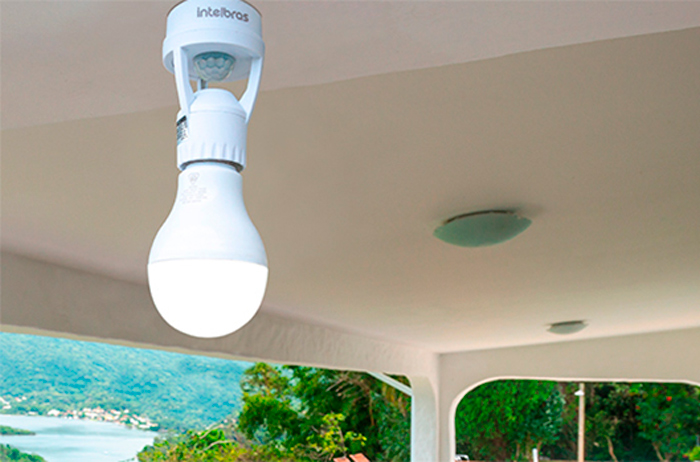 A imagem mostra um soquete adaptador com um sensor de presença integrado, fixado no teto. Uma lâmpada LED branca está rosqueada no soquete. O dispositivo é da marca Intelbras e parece estar instalado em uma varanda ou área externa coberta, com uma vista de paisagem com água e vegetação ao fundo. 