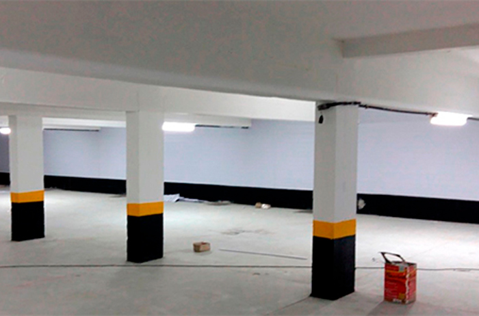 O ambiente é amplo e limpo, com paredes e teto brancos. O destaque são as colunas estruturais em cimento. A parte superior está pintada de branco, e a parte inferior, na altura do chão, está pintada com uma faixa de preto e uma faixa de amarelo vibrante logo acima, para sinalização de segurança.