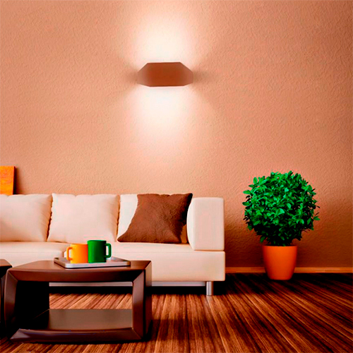 Esta arandela tem um design geométrico e discreto, com formato de um hexágono achatado e alongado na cor marrom. Ela é fixada rente à parede e emite luz indireta tanto para cima quanto para baixo, criando um efeito de iluminação suave e acolhedor em salas ou quartos.