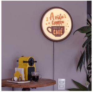 A placa redonda, com aro escuro e fundo claro iluminado, está pendurada na parede. Nela, lê-se a frase em português: "A vida começa depois do café" em letras laranjas e marrons, acompanhada de grãos de café e uma xícara. há uma máquina de café expresso