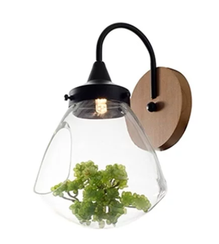 A imagem mostra uma arandela de parede com um design moderno. Possui uma base redonda de madeira clara fixada na parede. Dela, sai um braço curvo e preto que sustenta um globo de vidro transparente em formato de gota invertida.