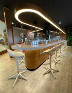Uma longa bancada de sushi em madeira clara, curva e iluminada por uma fita de LED suspensa no teto escuro, ocupa o centro. Em frente, há bancos altos brancos com bases em formato de estrela. Ao fundo, vê-se um letreiro "HIRO" e uma parede verde vertical de plantas.