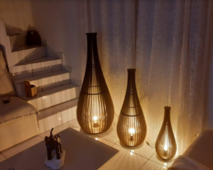 Três luminárias de chão, em formato de gota alongada e feitas de ripas de madeira clara, estão acesas, criando um brilho quente e padrões de sombra nas paredes e chão brancos. Elas variam em altura, do maior ao menor. No canto esquerdo, há uma escada branca e, em primeiro plano, a estatueta de elefante.