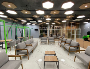 Uma moderna área de convivência interna com teto escuro e luminárias geométricas. O piso é branco e brilhante. Há vários sofás e poltronas cinzas com estrutura de madeira clara dispostos em grupos, e mesas de centro de metal e madeira. Ao fundo, estruturas verdes metálicas criam nichos.