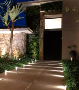 ma paisagem moderna ao anoitecer, mostrando um caminho largo de lajes de pedra cinza que leva a uma casa branca. O jardim lateral é decorado com plantas ornamentais e pedras, e é iluminado por pequenos balizadores com luz verde, destacando a vegetação. Ao fundo, há um lago e uma colina.