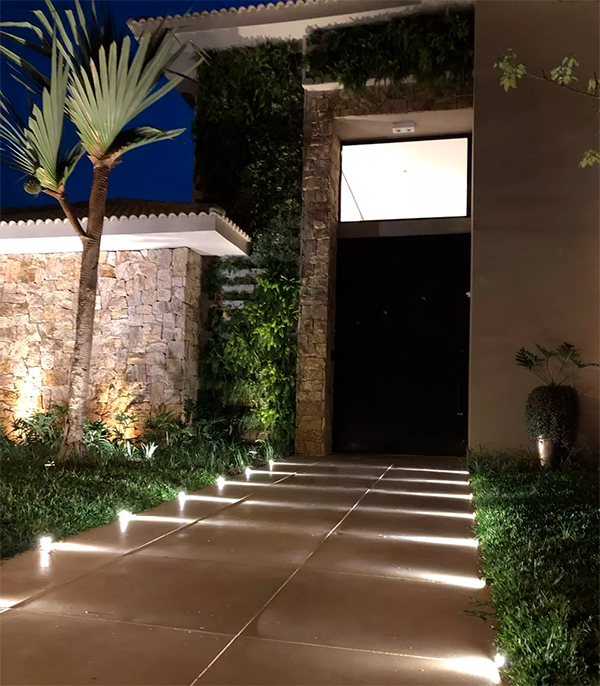 ma paisagem moderna ao anoitecer, mostrando um caminho largo de lajes de pedra cinza que leva a uma casa branca. O jardim lateral é decorado com plantas ornamentais e pedras, e é iluminado por pequenos balizadores com luz verde, destacando a vegetação. Ao fundo, há um lago e uma colina.