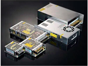 grupo de fontes de alimentação ou drivers metálicos, de formato retangular, dispostos sobre uma superfície escura e reflexiva. Os dispositivos possuem um acabamento prateado ou cinza. Têm tampas ou grades vazadas para ventilação. Há terminais de conexão expostos em uma das extremidades.